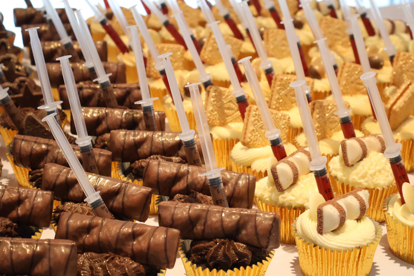 Rows of gold-cased cupcakes topped with chocolate bars and toy syringes of red sauce